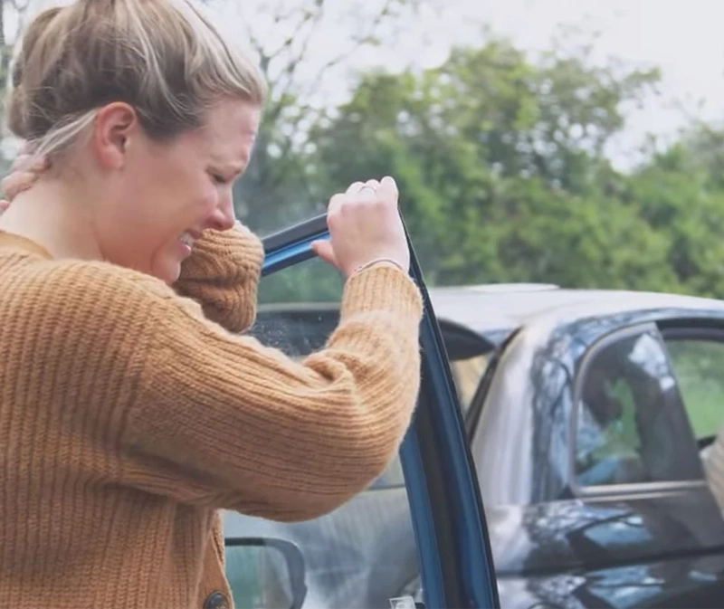 Blond woman winces in pain grasping her neck as she gets out of her car after having just been in a car accident